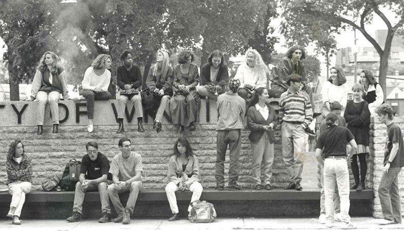 Black and white photo of students sitting in UWinnipeg sign